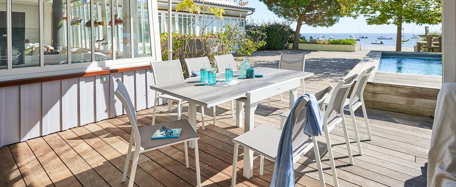 Terrasse extérieure avec table et chaises blanches, donnant sur une piscine et une vue panoramique. Les portes vitrées mènent à l'espace intérieur. Ensemble de salle à manger turquoise sur table.