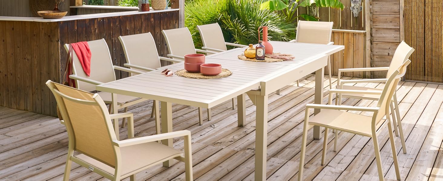 sur une terrasse en bois avec table rectangulaire, six chaises et un bar tiki. Plantes tropicales et ciel bleu visibles en arrière-plan.